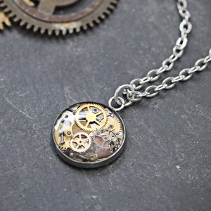 May include: A silver-toned necklace featuring a circular pendant filled with an assortment of small brass and silver-coloured metal gears. The pendant is attached to a simple silver-toned chain link necklace, displayed alongside several loose metal gears.
