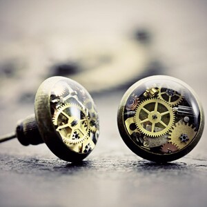 以下が含まれることがあります： ブロンズ調の金属製セッティングと透明なドーム型トップを備えた、円形のスタッドピアス一対。各ピアスの内部には、小さく精巧な金と銀の機械式時計の歯車やコグが層状に配置されているのが見えます。