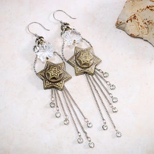 May include: A pair of dangle earrings featuring a clear faceted star-shaped bead at the top, connected to a larger antique gold-toned star charm with an embossed centre. Multiple silver-toned chains hang from the star, each tipped with a small clear crystal.
