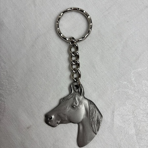 May include: A silver-toned metal keychain featuring a detailed horse head charm in profile. The charm is attached to a standard circular split ring by a short metal chain. The horse design shows a defined mane, ear, and facial features in a matte finish.