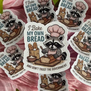 May include: A die-cut sticker featuring a cartoon raccoon wearing a pink chef hat and apron, slicing a loaf of bread on a wooden board. Next to the raccoon is a paper bag of bread and a bowl of flour. The text reads, I BAKE MY OWN BREAD AND DON'T TRUST THE GOVERNMENT.