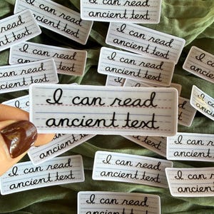 May include: A rectangular sticker featuring white paper with blue horizontal lines and a red baseline, printed with the black cursive text I can read ancient text. The sticker is shown in a collection of identical pieces.