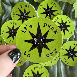 May include: A collection of circular lime green stickers featuring a black star graphic with a sad face and the text "it's punk to cry" printed in black lettering around the central star design. The stickers have a speckled texture.