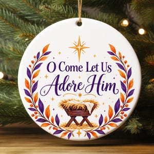 May include: A round white ceramic ornament featuring the text O Come Let Us Adore Him in purple script and serif font. The design includes a central wooden manger with straw, a gold star above, and a wreath of purple and orange leaves.