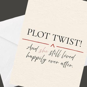 Puede incluir: Una tarjeta de felicitación de color crema con un sobre a juego. La tarjeta presenta el texto negro PLOT TWIST! sobre una fina línea horizontal roja y un pequeño cheurón rojo. Debajo, el texto en cursiva negra dice And she still lived happily ever after.