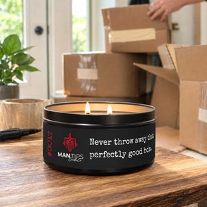 Può includere: Una candela in barattolo di metallo nero con due stoppini accesi. L'etichetta presenta un logo rosso, il testo #0017 e le parole MAN TIPS Candle Co. seguite dal testo: Never throw away that perfectly good box. La candela è su legno.