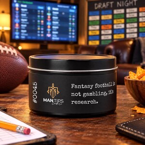 May include: A black cylindrical candle tin with a matte finish. The label features the text #0045, the Man Tips Candle Co logo in gold, and the white printed text: Fantasy football is not gambling, it's research. The lid is also black.