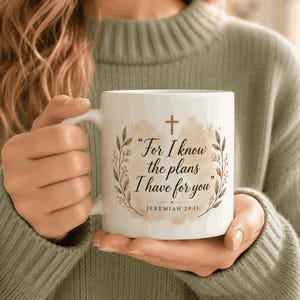 May include: A white ceramic mug featuring a brown watercolor-style background with a cross, leafy branches, and the text: For I know the plans I have for you, Jeremiah 29:11. The design is centered on the front of the mug with a handle on the left side.