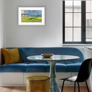 May include: A framed landscape painting featuring a vibrant green and yellow field under a dramatic blue sky with white clouds, displayed on a light gray wall above a blue velvet sofa, a round pedestal table, and a mustard yellow ottoman.