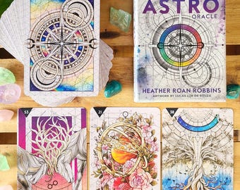 May include: A 56-card deck and guidebook for the Starcodes Astro Oracle. The cards feature colorful illustrations with celestial themes, including a compass, a tree, and a celestial sphere. The guidebook is written by Heather Roan Robbins and features artwork by Lucas Lua de Souza.