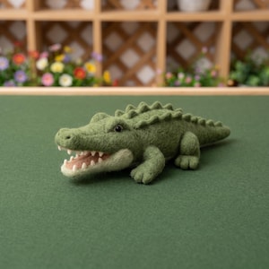 May include: A needle-felted crocodile figurine in shades of olive and sage green. The creature features a textured body, a row of dorsal scales along its back, a light cream-coloured underbelly, and an open mouth revealing small white felt teeth.