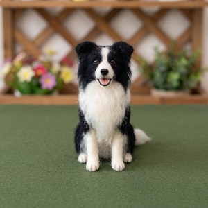 May include: A needle-felted sculpture of a Border Collie dog sitting in an upright position. The figure features black and white fur, a white chest, a black nose, and an open mouth showing a pink tongue. The texture appears soft and fuzzy.