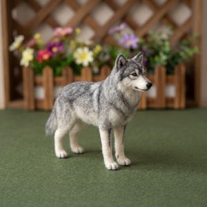 May include: A needle-felted wolf figurine featuring a realistic coat of gray, white, and black fibers. The wolf stands in a profile position with pointed ears, alert eyes, and a black nose, showcasing detailed texture throughout its body.
