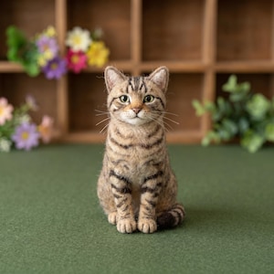 May include: A needle-felted cat figurine featuring a tan and brown tabby coat with dark stripes, pointed ears, and green eyes. The cat is depicted in a seated position with its tail curled around its side, showcasing a textured, soft fibre finish.