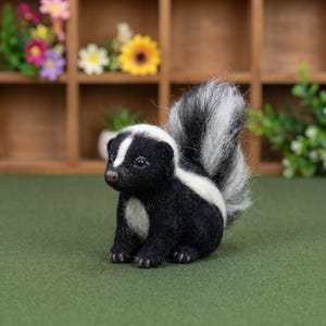 May include: A needle-felted skunk figurine featuring a black body with a white stripe running from the top of its head down its back. The skunk has a fluffy, upright tail with grey and white fur, small dark eyes, and a brown nose, sitting upright.