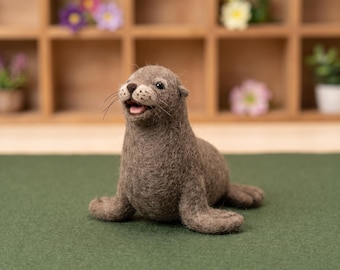 Op de afbeelding: Een naaldgevilte bruine zeehondfiguur met een textuur van wol, donkere ogen, een kleine zwarte neus en een geopende mond met een roze tong. De zeehond heeft snorharen en de voorvinnen zijn naar voren gericht in een zittende houding.