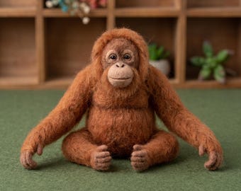 May include: A needle-felted orangutan figurine with textured, reddish-brown wool fur. The figure has a rounded face, dark eyes, and a light brown muzzle, sitting in a forward-facing position with long arms extended outward on a green surface.
