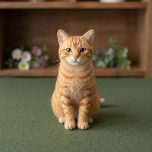Könnte beinhalten: Eine nadelgefilzte orangefarbene getigerte Katzenfigur mit ausgeprägten dunkelorangen Streifen, spitzen Ohren und bernsteinfarbenen Augen. Die Katze ist in sitzender Position dargestellt und zeigt eine strukturierte Oberfläche aus Wolle.
