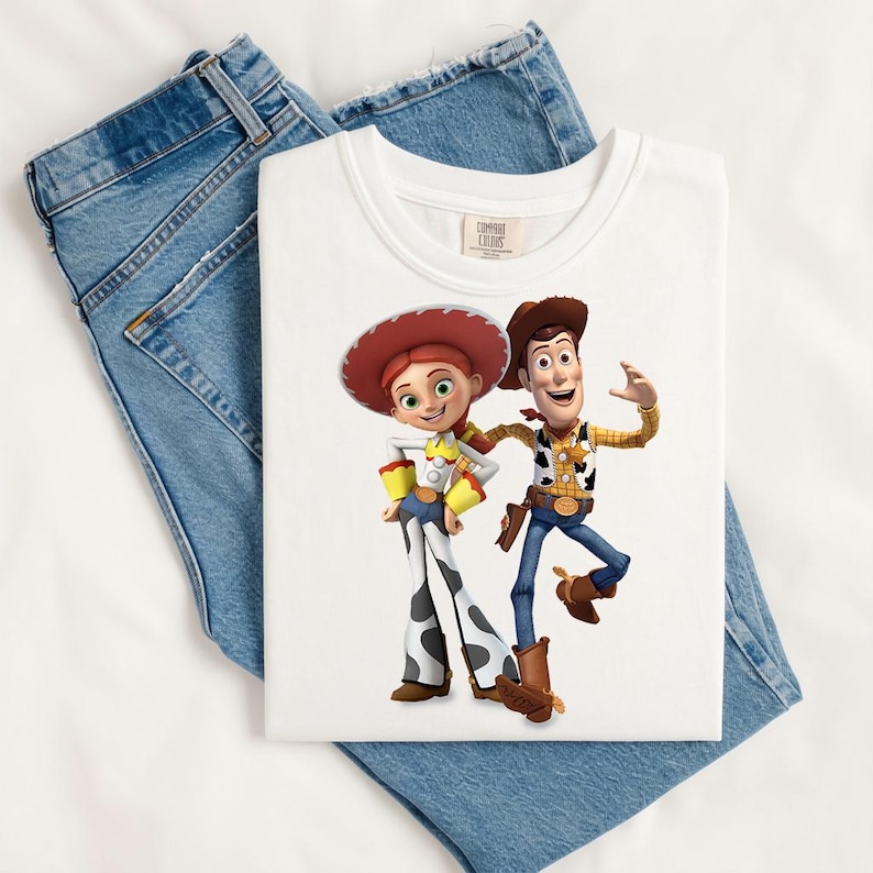 May include: A white crew neck t-shirt featuring a graphic print of the characters Jessie and Woody from Toy Story. Jessie wears a red cowgirl hat, yellow shirt, and cow-print chaps. Woody wears a brown cowboy hat, yellow shirt, and blue jeans.