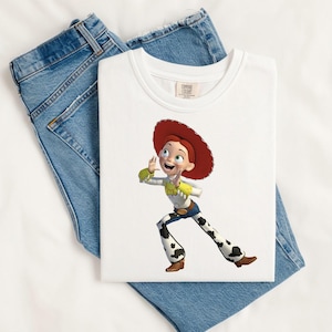 May include: A white crew neck t-shirt featuring a graphic print of the character Jessie from Toy Story, who wears a red cowgirl hat, a yellow and white shirt, and white pants with black cow-print spots, layered over a pair of blue denim jeans.