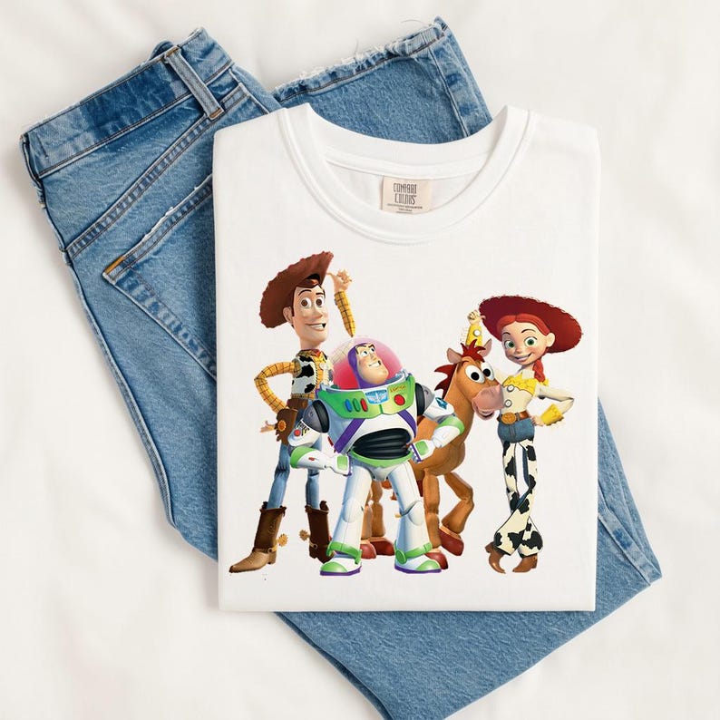 May include: A white t-shirt featuring a graphic print of the Toy Story characters Woody, Buzz Lightyear, Bullseye, and Jessie. The shirt is layered over a pair of blue denim jeans. The graphic shows the characters standing together in full color.