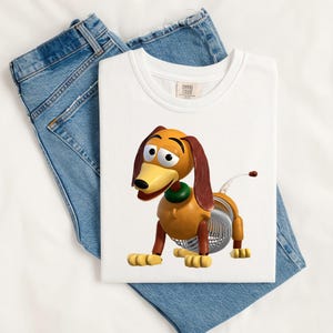 May include: A white t-shirt featuring a graphic of a brown and tan toy dachshund with long floppy ears, a green collar, and a silver metal spring body. The shirt is displayed alongside a pair of blue denim jeans with visible stitching.