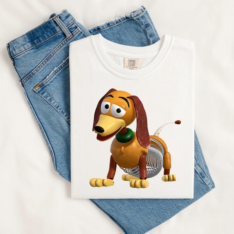 May include: A white t-shirt featuring a graphic of a brown and tan toy dachshund with long floppy ears, a green collar, and a silver metal spring body. The shirt is displayed alongside a pair of blue denim jeans with visible stitching.