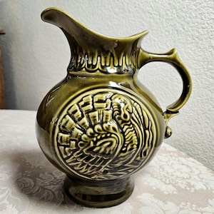 May include: A glossy olive green ceramic pitcher featuring a rounded body with a prominent embossed circular medallion depicting a stylized turkey. The vessel includes a curved handle, a flared spout, and a decorative scalloped rim detail.