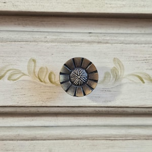 May include: A round, dark bronze-toned metal drawer knob with a sunburst pattern, featuring a central textured circle surrounded by radiating spokes, mounted on a light-colored wood drawer front with painted green leaf accents.