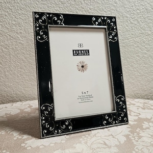 May include: A rectangular picture frame with a glossy black border featuring white scrollwork and dot patterns. The frame has a silver beaded inner and outer edge. The insert displays the text Burnes of Boston, 5 x 7, and a flower graphic.