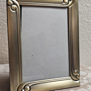 May include: A rectangular picture frame with a brushed antique gold finish. The frame features a smooth, metallic surface with decorative, raised scrollwork details at each of the four corners. A plain, light gray insert fills the center area.