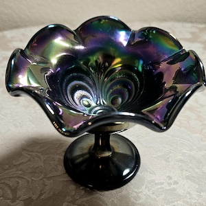 May include: A dark purple carnival glass bowl with an iridescent finish and a ruffled, scalloped edge. The bowl features a molded pattern of stylized peacock feathers on the interior and is supported by a solid, circular pedestal base.