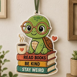 May include: A hanging embroidered wall decoration featuring a green turtle wearing orange glasses, sitting on a stack of three books. The books are labelled READ BOOKS, BE KIND, and STAY WEIRD. A steaming mug with a heart and a small heart icon are included.
