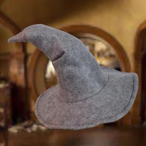 Puede incluir: Un sombrero puntiagudo de fieltro gris con un ala ancha y ligeramente curvada. El sombrero presenta una copa cónica alta que se estrecha hacia una punta suave y redondeada. El material tiene una apariencia texturizada y jaspeada en gris.