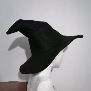 Könnte beinhalten: Ein einfarbiger, schwarzer Spitzhut mit einer breiten, flexiblen Krempe. Der Hut hat eine hohe, leicht gebogene Krone, die spitz zuläuft. Das Material wirkt weich und filzartig, und die Krempe weist eine sanfte, unebene Wölbung am Rand auf.