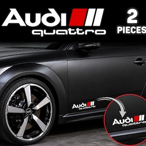 May include: A set of two Audi quattro vinyl decals featuring white text and a red square graphic. The decals are shown applied to the lower side panel of a matte black car, with a circular inset highlighting the placement of the sticker.