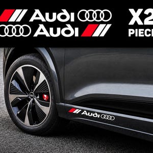 Könnte beinhalten: Ein Set aus zwei weißen Vinyl-Aufklebern mit dem Audi-Logo und Schriftzug sowie einem schrägen rot-schwarzen Streifenmuster. Die Aufkleber sind an der unteren Seitenverkleidung eines dunkelgrauen Fahrzeugs angebracht.
