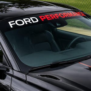 Op de afbeelding: Een zwarte autoruit voorzien van een horizontale zonwerende sticker met de tekst FORD PERFORMANCE in witte en rode hoofdletters. De sticker is aangebracht op het bovenste gedeelte van het glas, boven de achteruitkijkspiegel en het dashboard.