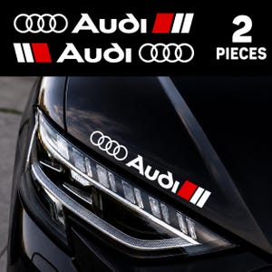 以下が含まれることがあります： Audiのロゴとテキスト、および赤と白の長方形のアクセントブロックがデザインされた2枚の白いビニール製デカール。車の黒いボンネットに貼られており、ヘッドライトユニットの上部に斜めに配置されています。