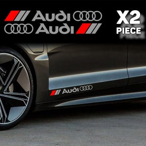 以下が含まれることがあります： 赤とグレーの斜めストライプデザインと、Audiのブランド名、および白い4つのリングロゴが特徴のAudi車用デカール2枚セット。デカールは、ダークグレーの車両のフロントホイール近くの下部サイドパネルに貼り付けられた状態で表示されています。