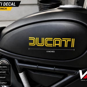 May include: A yellow vinyl decal featuring the word DUCATI in a stylised font applied to a matte black motorcycle fuel tank. The decal measures 15.24 cm in length as indicated by a graphic line and text below the lettering on the tank surface.