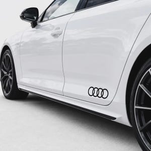 Peut inclure: Une berline blanche arborant un autocollant noir représentant le logo aux quatre anneaux d'Audi sur la partie inférieure de la portière passager. Le véhicule est équipé de jantes en alliage sombres, d'un rétroviseur noir et d'une poignée noire.
