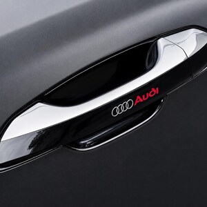 May include: A sleek black car door handle featuring a polished chrome accent strip along the top edge. The Audi logo and the word Audi are printed in red and white on the handle surface, set against a matte black car body panel finish.