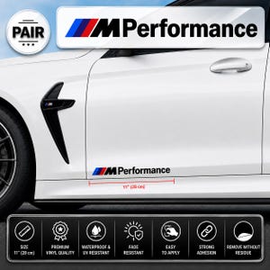 Op de afbeelding: Een wit zijpaneel van een auto met een zwarte M Performance sticker met blauwe, paarse en rode strepen. De sticker is 28 cm lang. Een cirkelvormig embleem geeft aan dat een paar is inbegrepen en een paneel onderaan somt de kenmerken op.