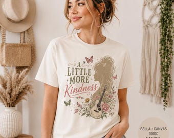 May include: A natural-colored t-shirt featuring a graphic print with the text A LITTLE MORE KINDNESS, a silhouette of a person with long hair, a guitar, butterflies, and floral elements in shades of green, pink, and brown on the front.