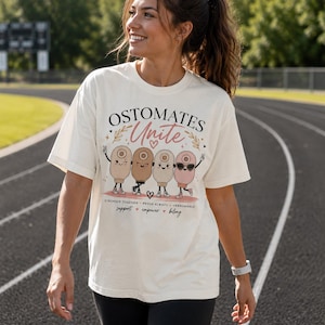 May include: An off-white short-sleeve t-shirt featuring a graphic of four cartoon ostomy bags with faces, arms, and legs. The text reads OSTOMATES Unite, STRONGER TOGETHER PROUD ALWAYS UNBREAKABLE, and support empower belong with small heart icons.
