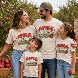 Puede incluir: Cuatro personas con camisetas de manga corta a juego en color crema con el texto en rojo y verde "APPLE PICKING CREW" y un pequeño gráfico de una cesta de manzanas. Las camisetas tienen una fuente retro y están en un huerto de manzanas.