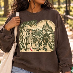 May include: A dark brown crewneck sweatshirt featuring a graphic print of the Tin Man, Dorothy, the Cowardly Lion, and the Scarecrow walking on a yellow brick road toward the Emerald City, with Dorothy wearing bright red shoes.