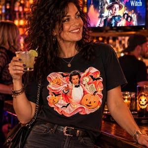 May include: A black short-sleeved t-shirt featuring a graphic print of a person in a tuxedo cape surrounded by orange jack-o'-lanterns, a skeleton, and black bats against a pink fabric background. The shirt is worn with dark blue denim jeans.
