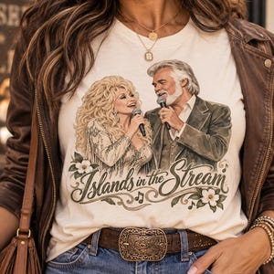 Puede incluir: Una camiseta de color crema con una ilustración gráfica de una mujer rubia y un hombre con barba gris cantando en micrófonos. Debajo de las figuras, el texto Islands in the Stream está impreso en letra cursiva, decorado con motivos florales.